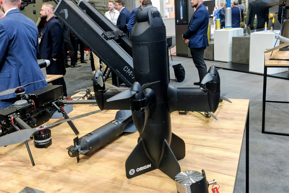 Czarny dron przechwytujący BLAZE firmy Origin Robotics, ustawiony pionowo na stole wystawowym, z czterema wirnikami wokół kadłuba.