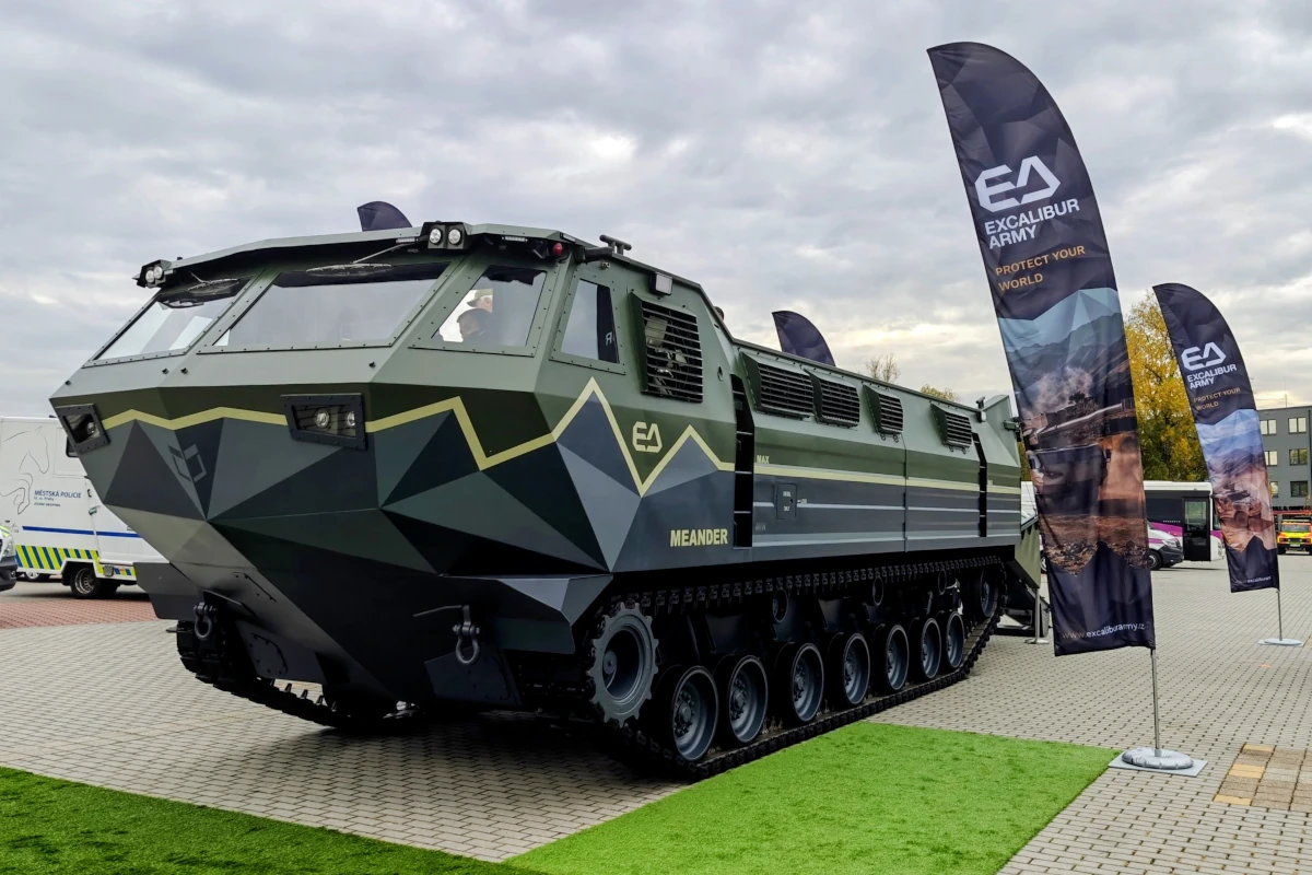 Gąsienicowy transporter amfibijny Meander w geometrycznym kamuflażu na wystawie plenerowej, obok flag Excalibur Army.