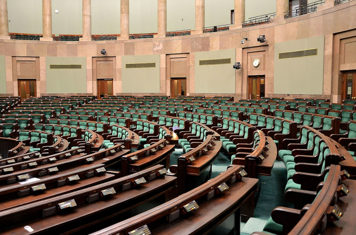 Pusta sala posiedzeń Sejmu Rzeczypospolitej Polskiej z rzędami zielonych foteli i pulpitami do głosowania
